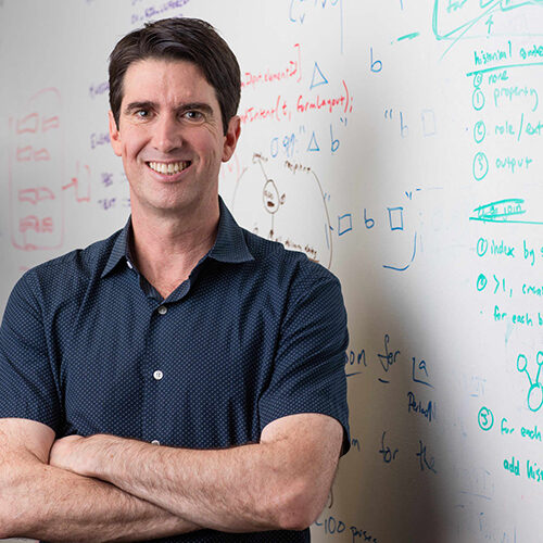 Portrait of Adam Cheyer, Creator of Siri standing in front of a whiteboard with text.