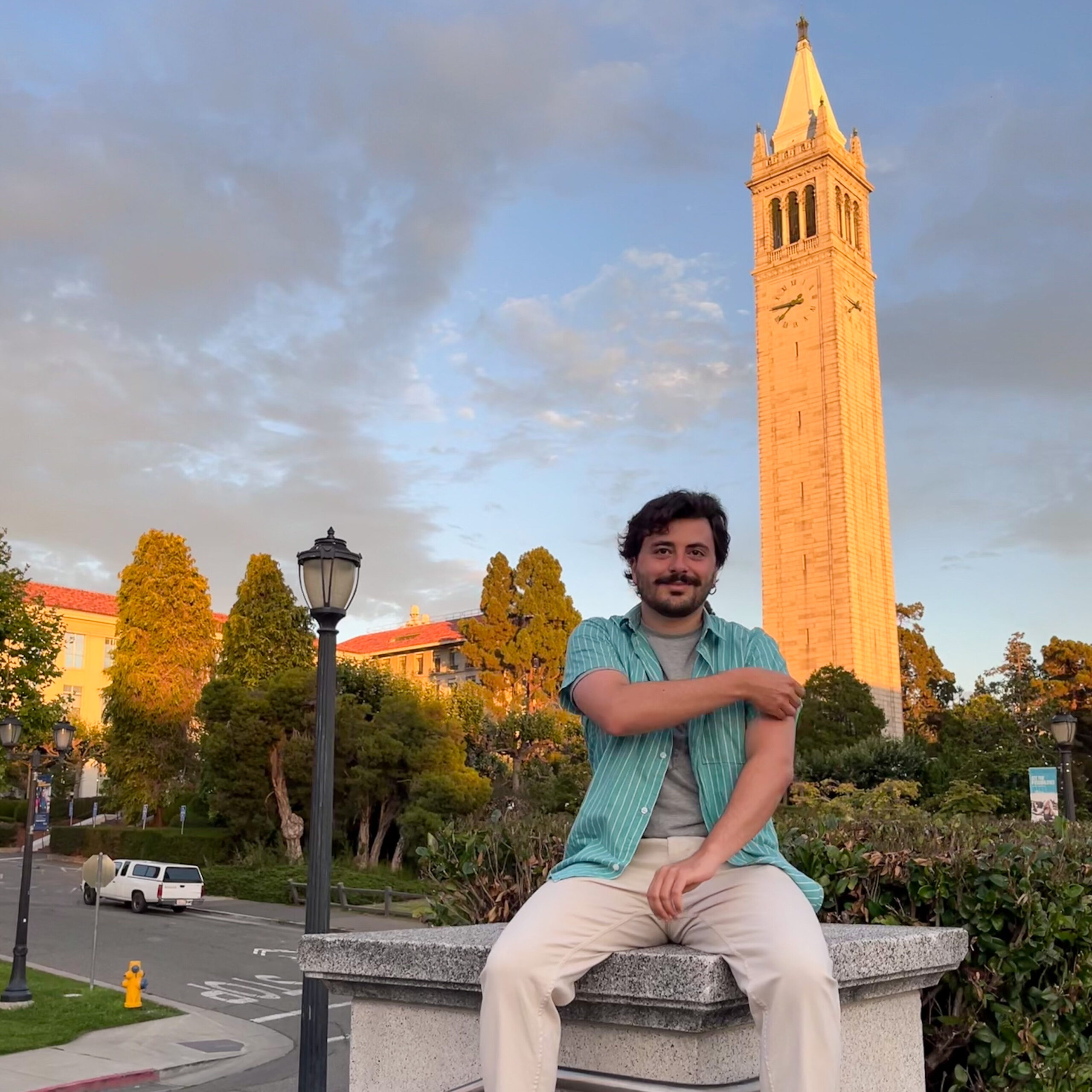 Giulio Pantano sits on a pillar with the Berkeley Campanile tower in the backdrop around sunset. 