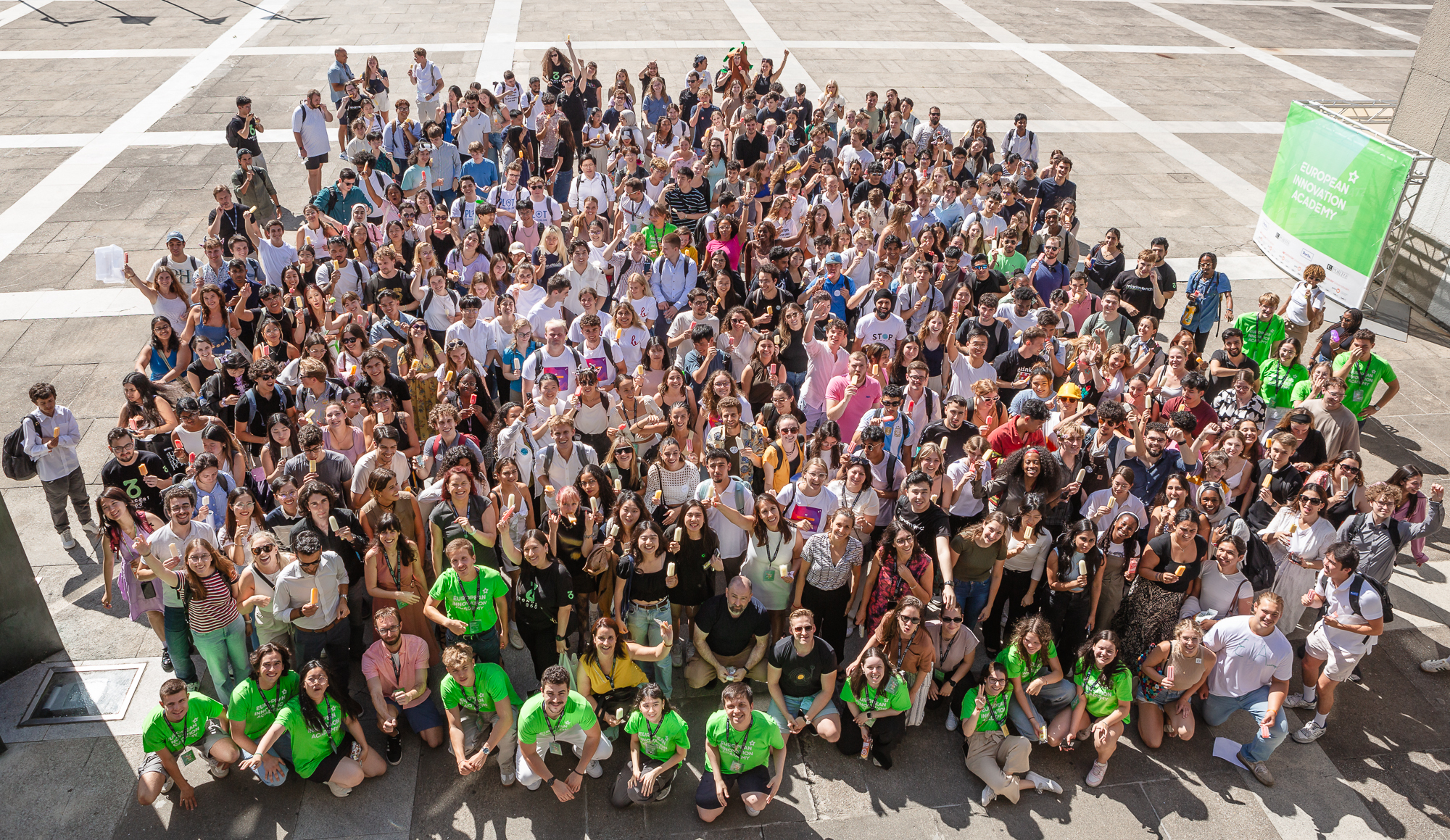 The participants from the European Innovation Academy in Porto pose for a group photo taken form a bird's-eye view. They stand closely together outdoors on a sunny day, smiling and facing the camera.