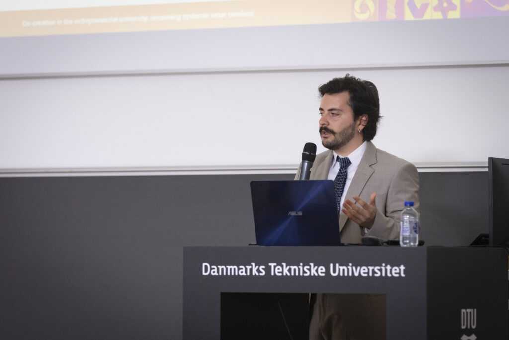 Giulio Pantano stands at the podium at Danmarks Tekniske Universitet as he presents his PhD defense in front of a gray backdrop