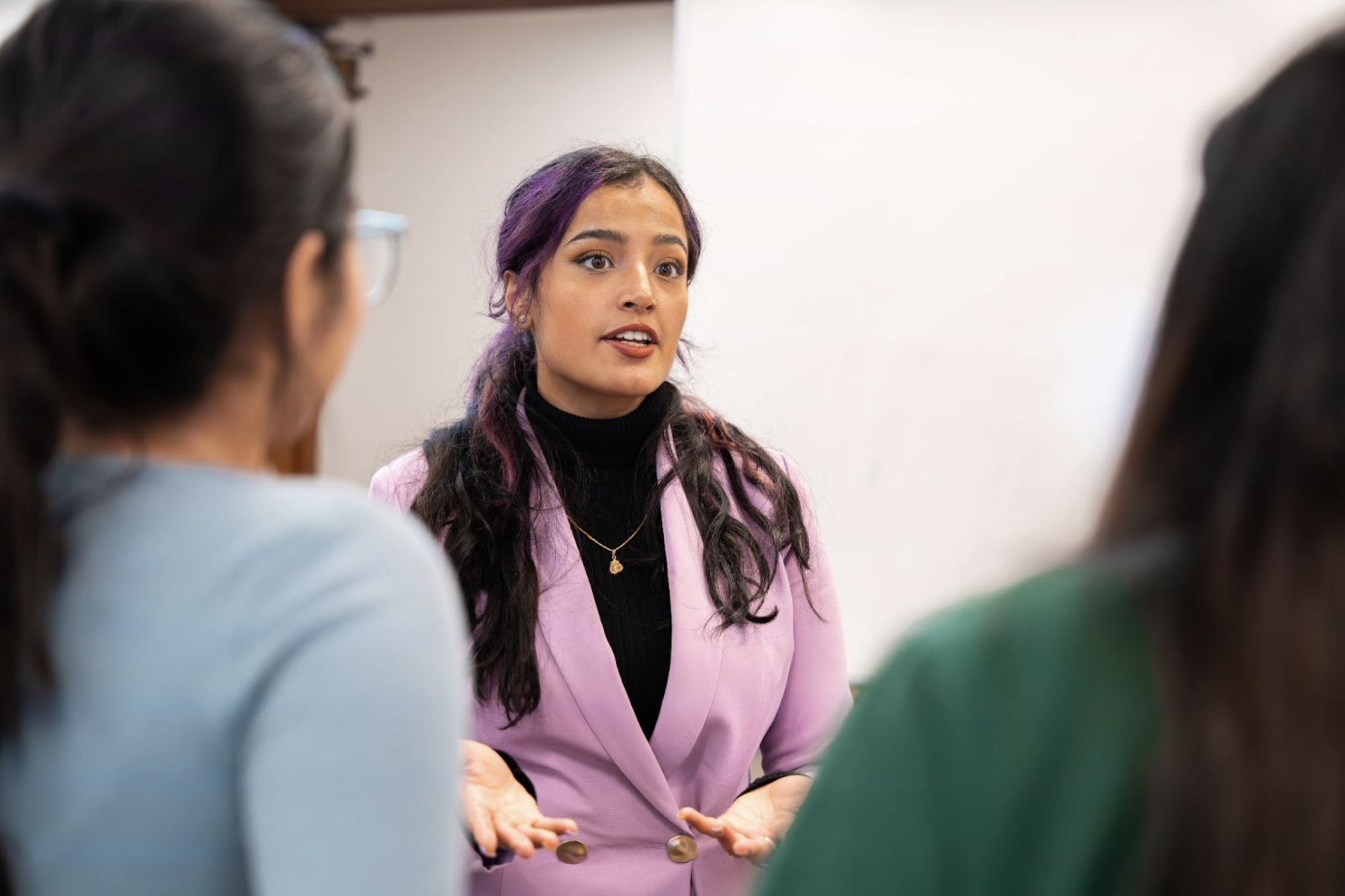 Kashish Juneja, a woman in a pink suit, stands centered and speaking in the direction of the camera. The backs of two students, are seen at the edges of the frame as they listen to Kashish.