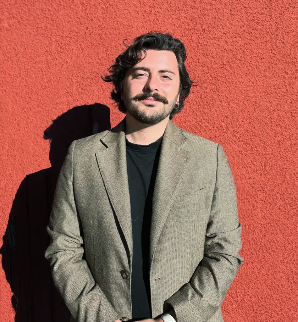 Giulio Pantano smiles for a headshot taken in direct sunlight against a bright red wall